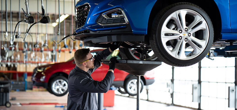 A mechanic in a black uniform and gloves performing maintenance on a car lifted up in an auto repair shop.
