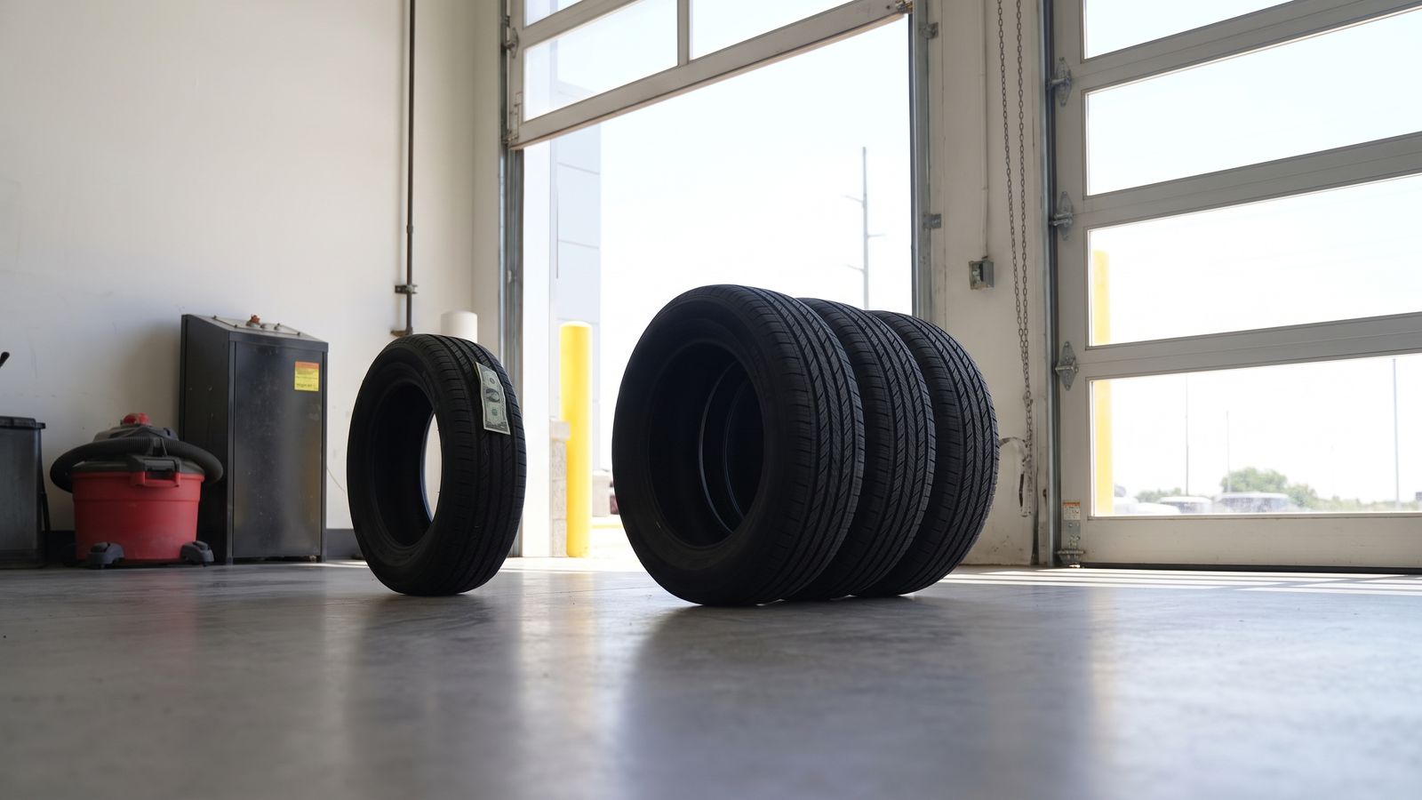 Three new tires huddled together on a Motor Inn service bay floor while a fourth tire, a dollar bill tucked in its tread, stands apart.