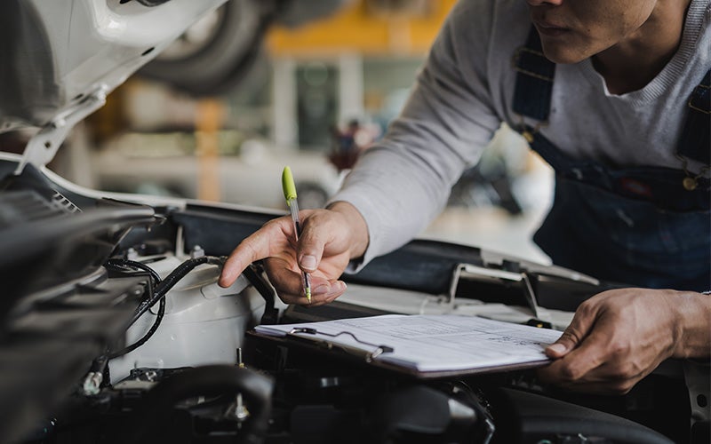A technician inspecting a car engine and writing on a clipboard.